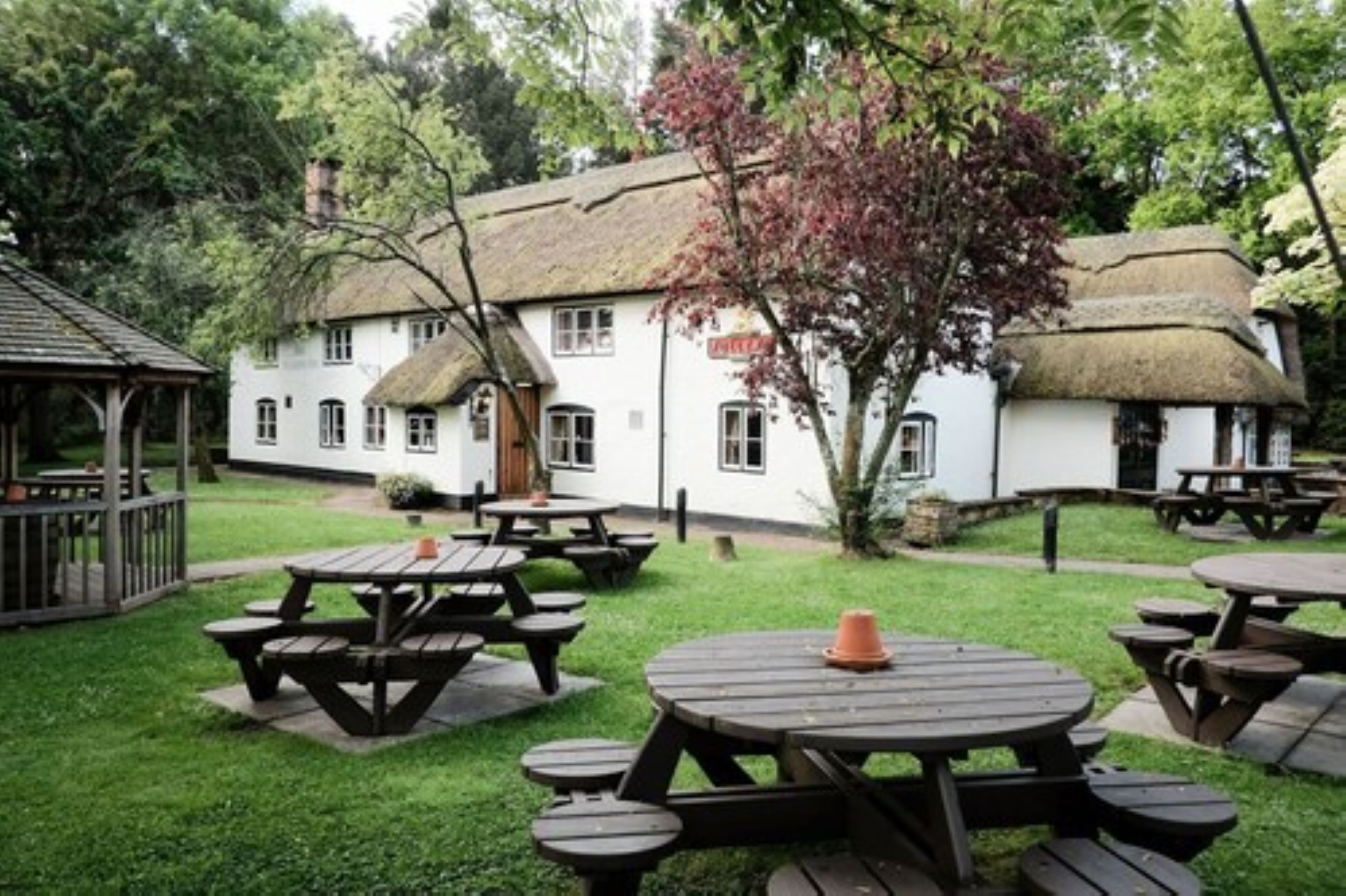 A rustic outdoor seating area with wooden tables and benches, surrounded by a white building with a thatched roof.