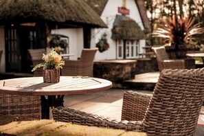 A round wooden table with a small potted plant, wicker chairs, and a thatched-roof building in the background.