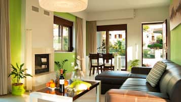 A living room with a sofa, a coffee table with fruit and a bottle, a fireplace, and a dining area with chairs and a table.