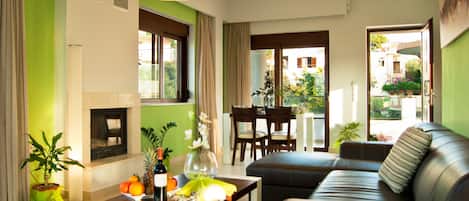 Una sala de estar con sofá, una mesa de centro con fruta y una botella, chimenea y un comedor con sillas y una mesa.