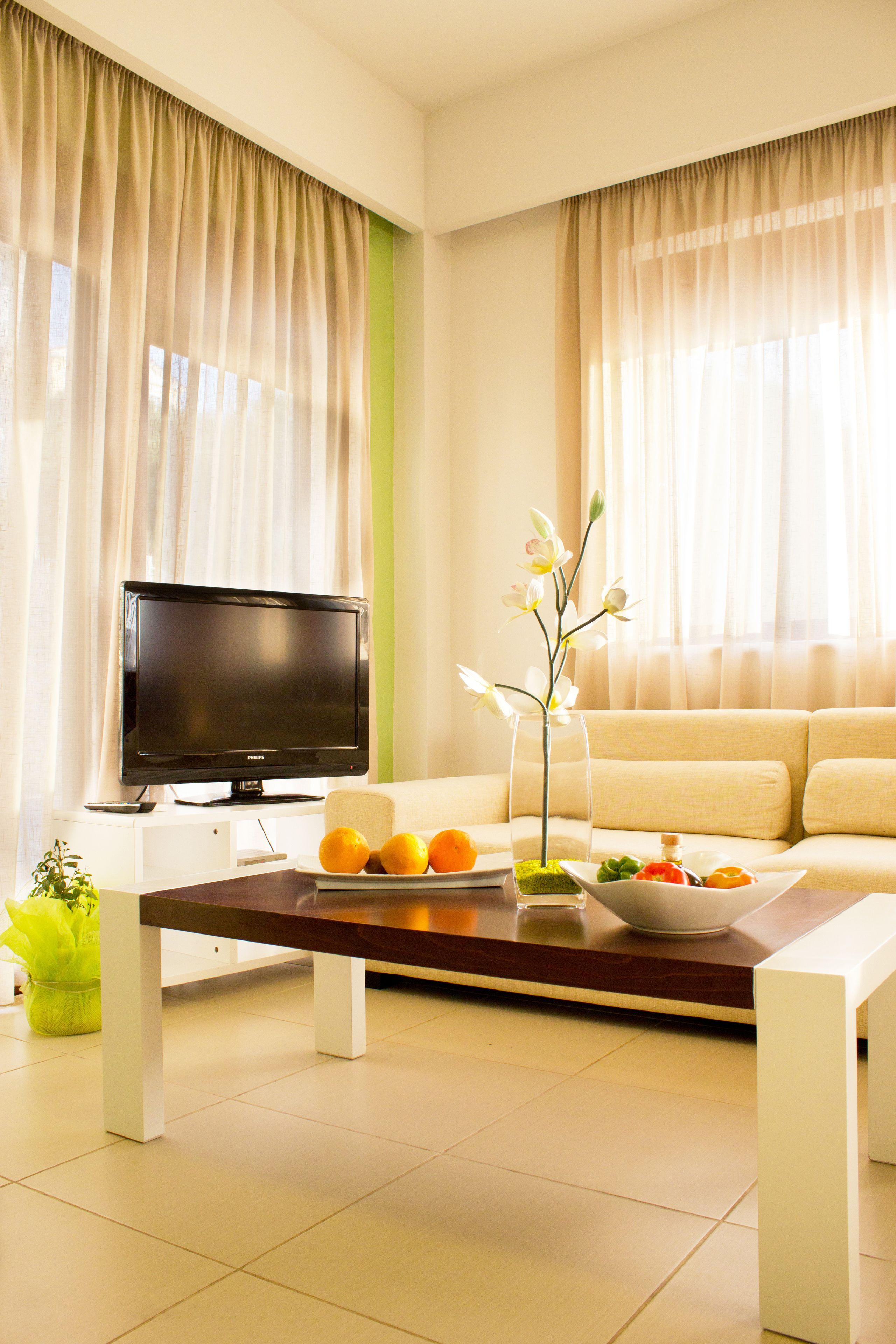 A modern living room with a flat-screen TV, a sofa, a coffee table with fruits and a vase, and sheer curtains.