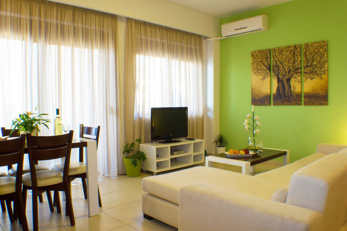 Una sala de estar con televisor, sofá, mesa de comedor, sillas y una planta en maceta.