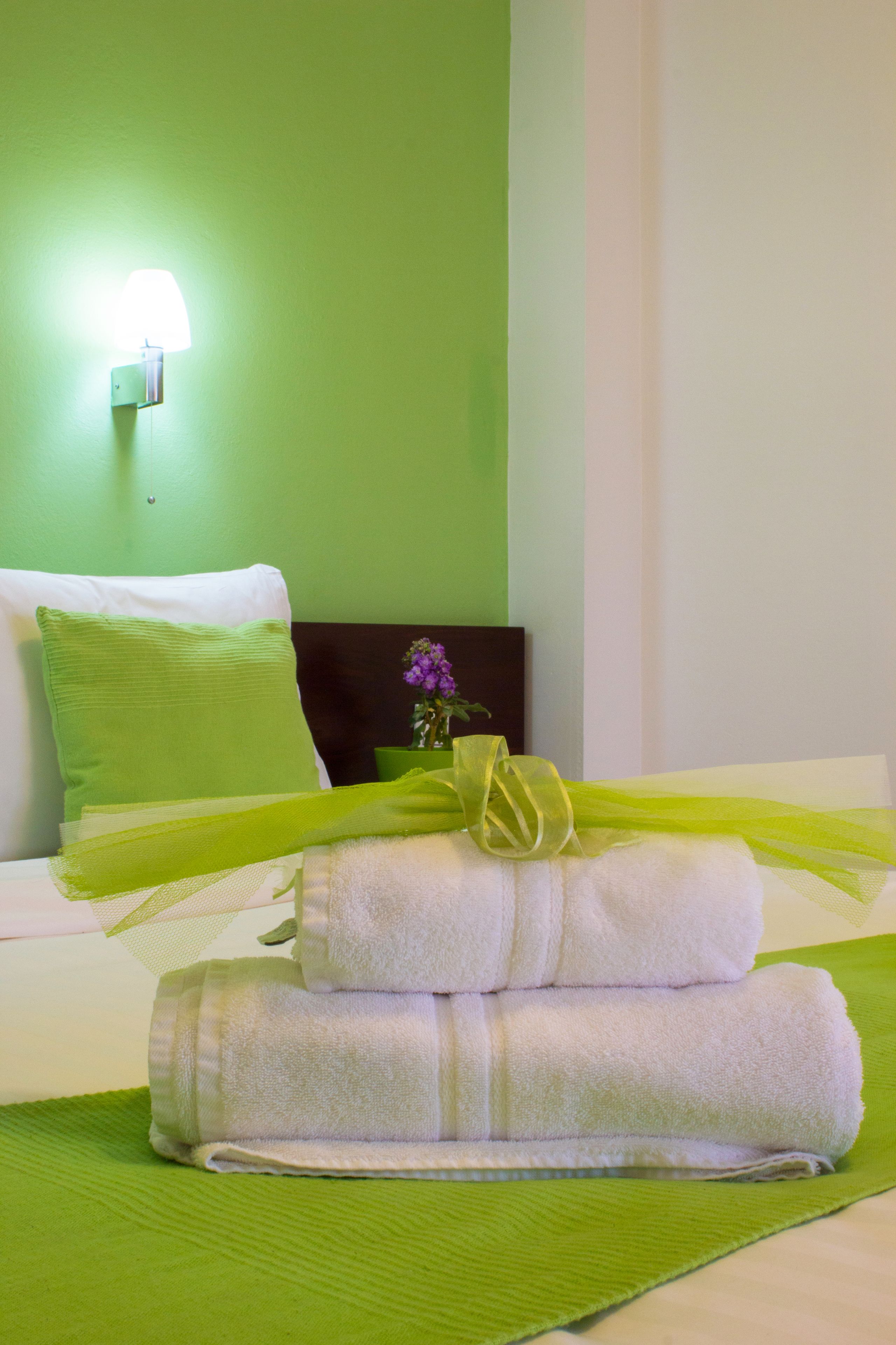A neatly made bed with green bedding and a white pillow, a vase with purple flowers, and a wall-mounted lamp.