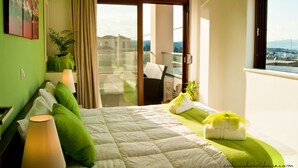 A hotel room with a bed, green pillows, a green plant, a window with a view, and a balcony with a chair.