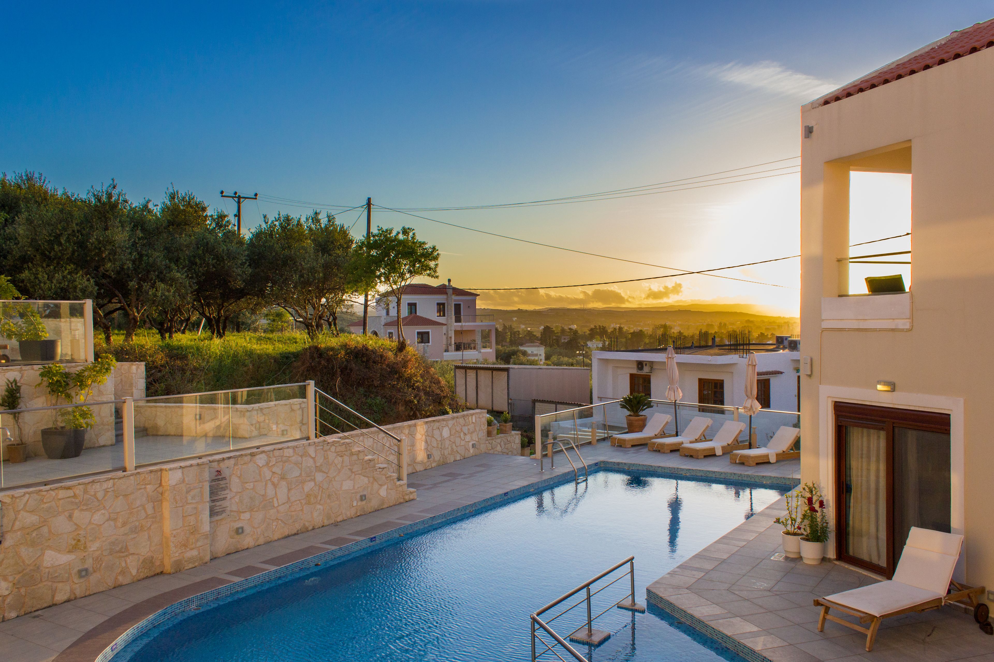 A swimming pool with lounge chairs and a stone wall, surrounded by greenery and buildings.