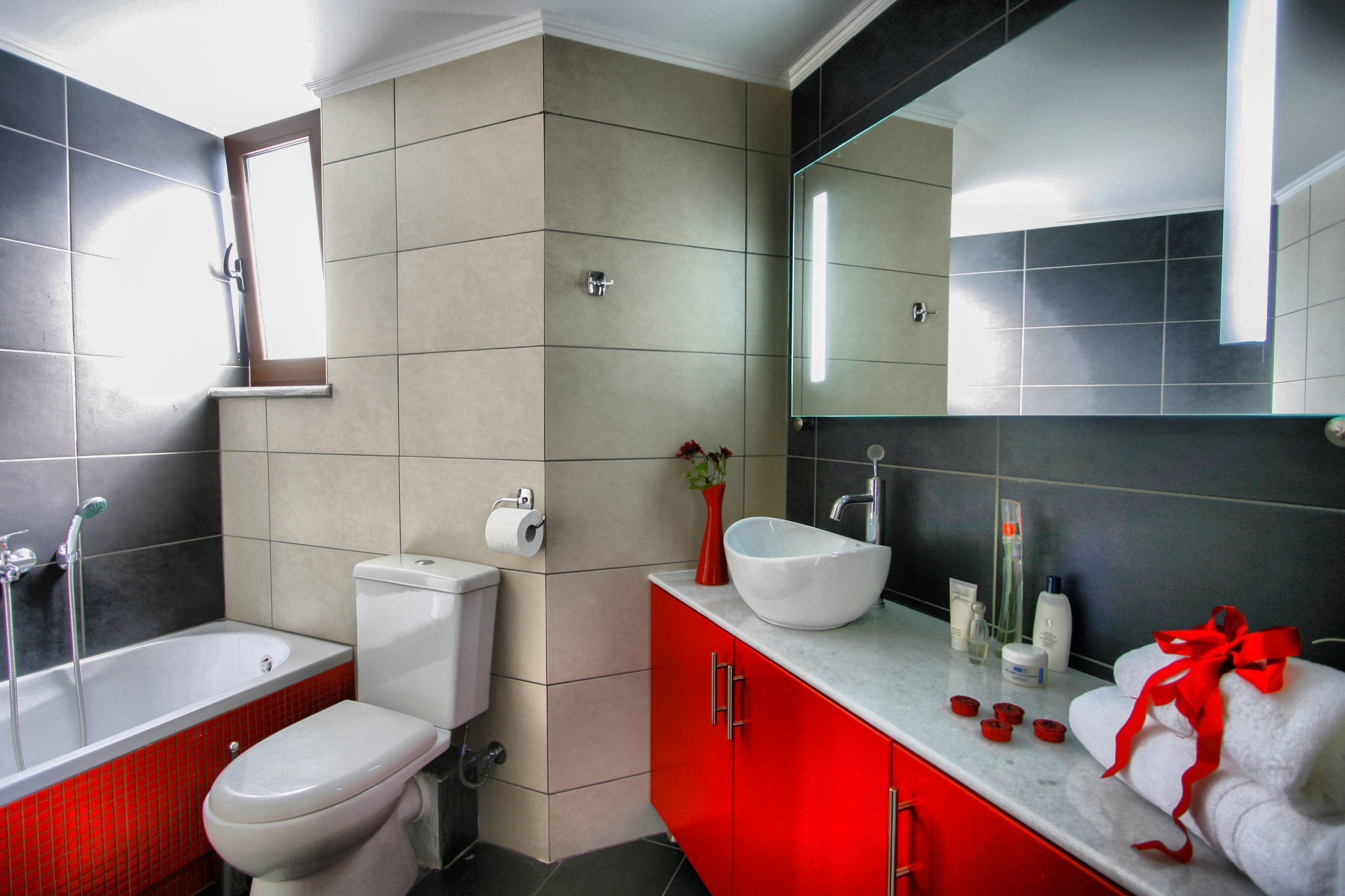 A modern bathroom with a bathtub, toilet, vanity with a large mirror, and a red cabinet.