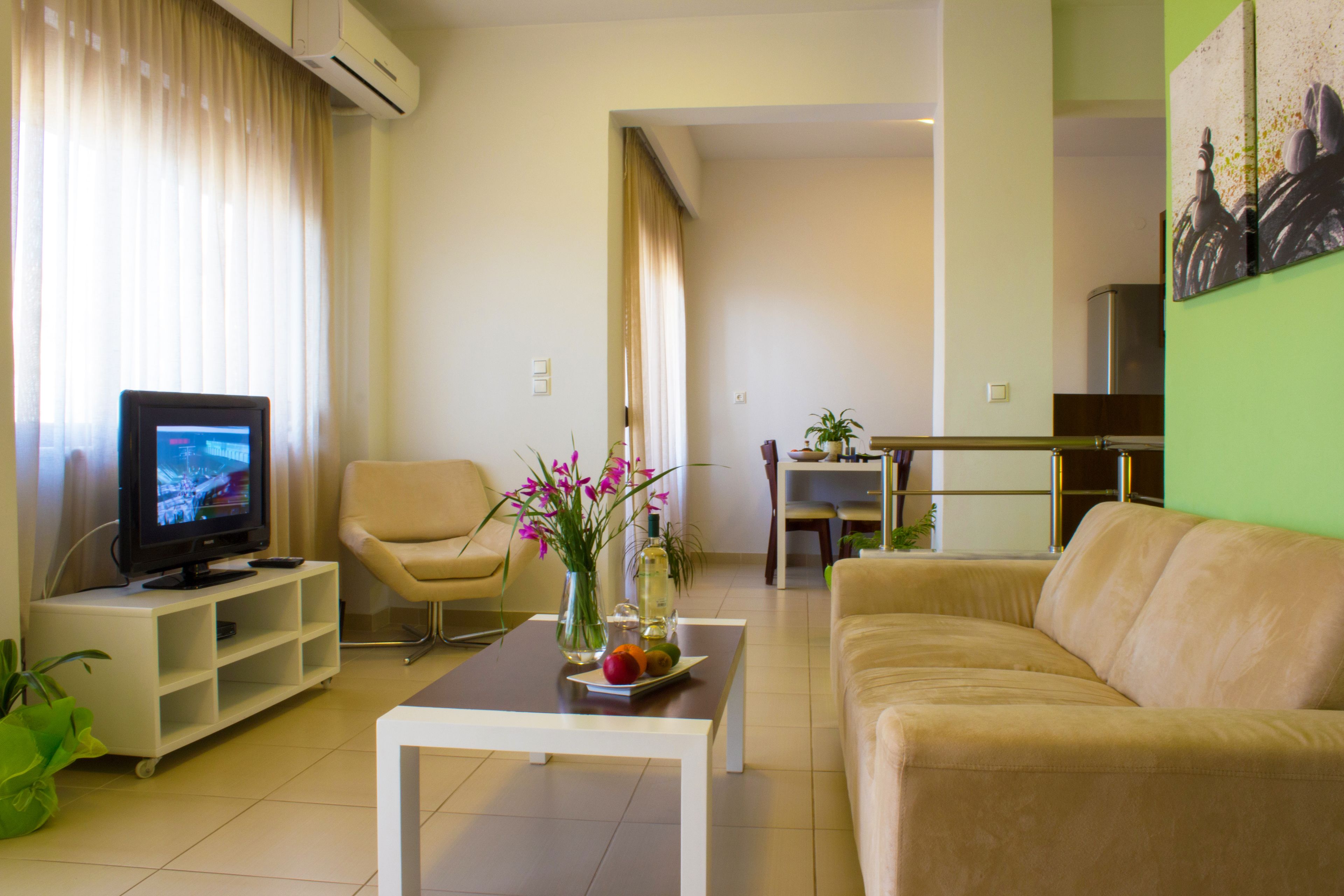 Uma sala de estar com televisão, um sofá bege, uma mesa de centro com um vaso de flores e uma poltrona.