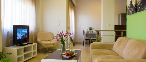 Una sala de estar con televisor, un sofá color beige, una mesa de centro con un jarrón de flores y una silla.