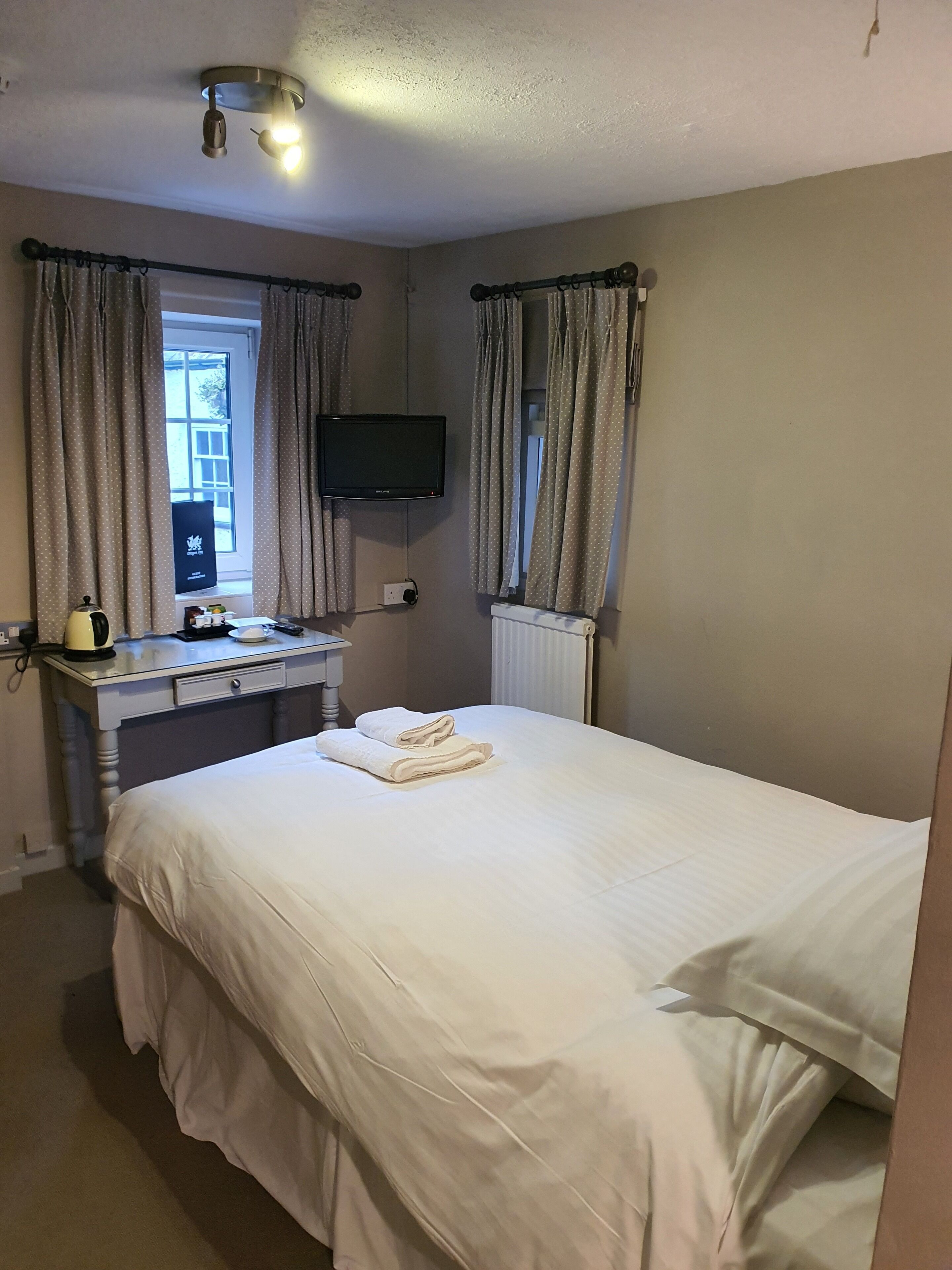 A hotel room with a bed, a desk with a kettle, a television, and curtains.