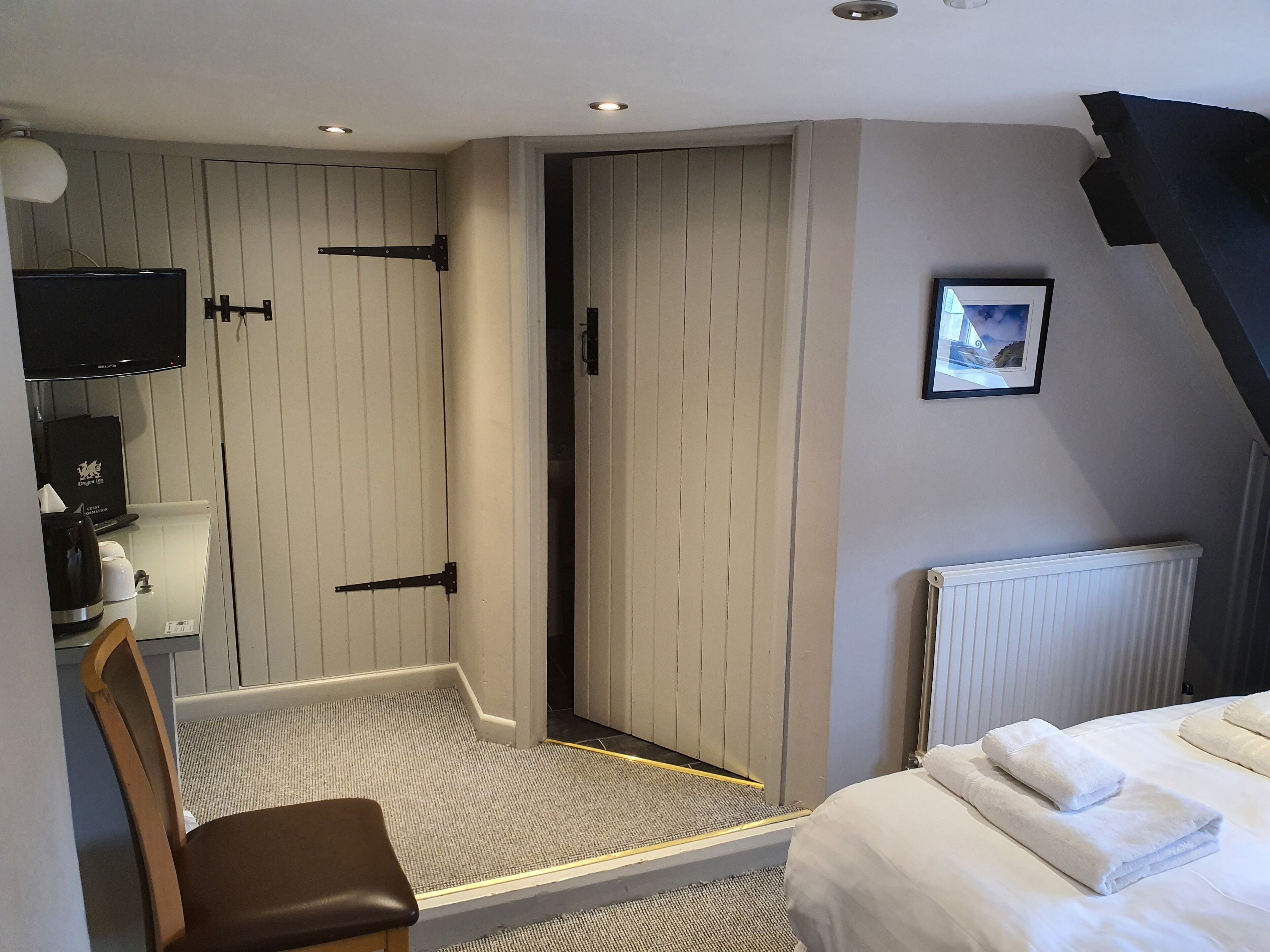 A hotel room with a bed, a chair, a shower area, a television, and a framed picture on the wall.