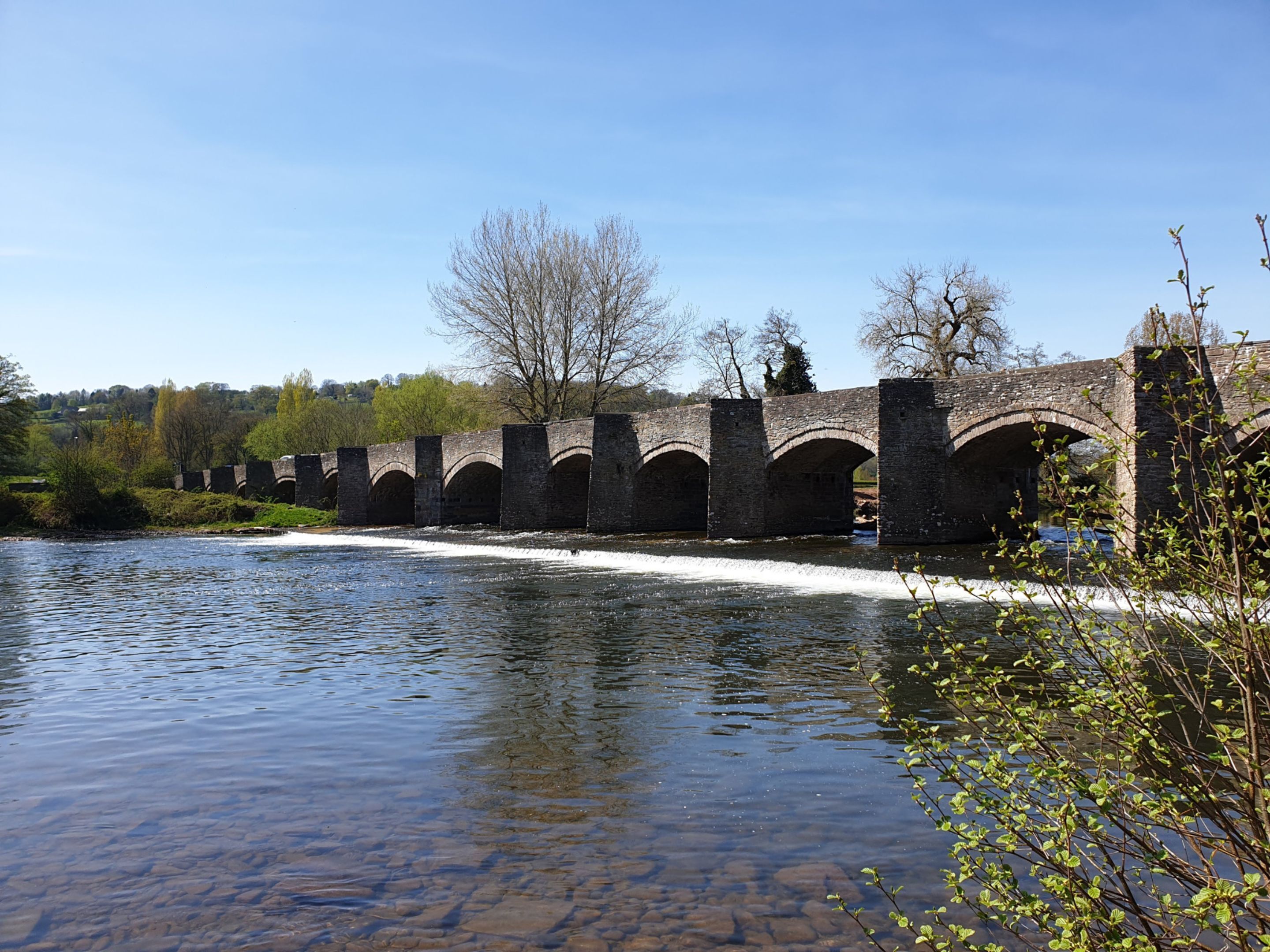 A stone bridge over a river with clear water, surrounded by trees and greenery.