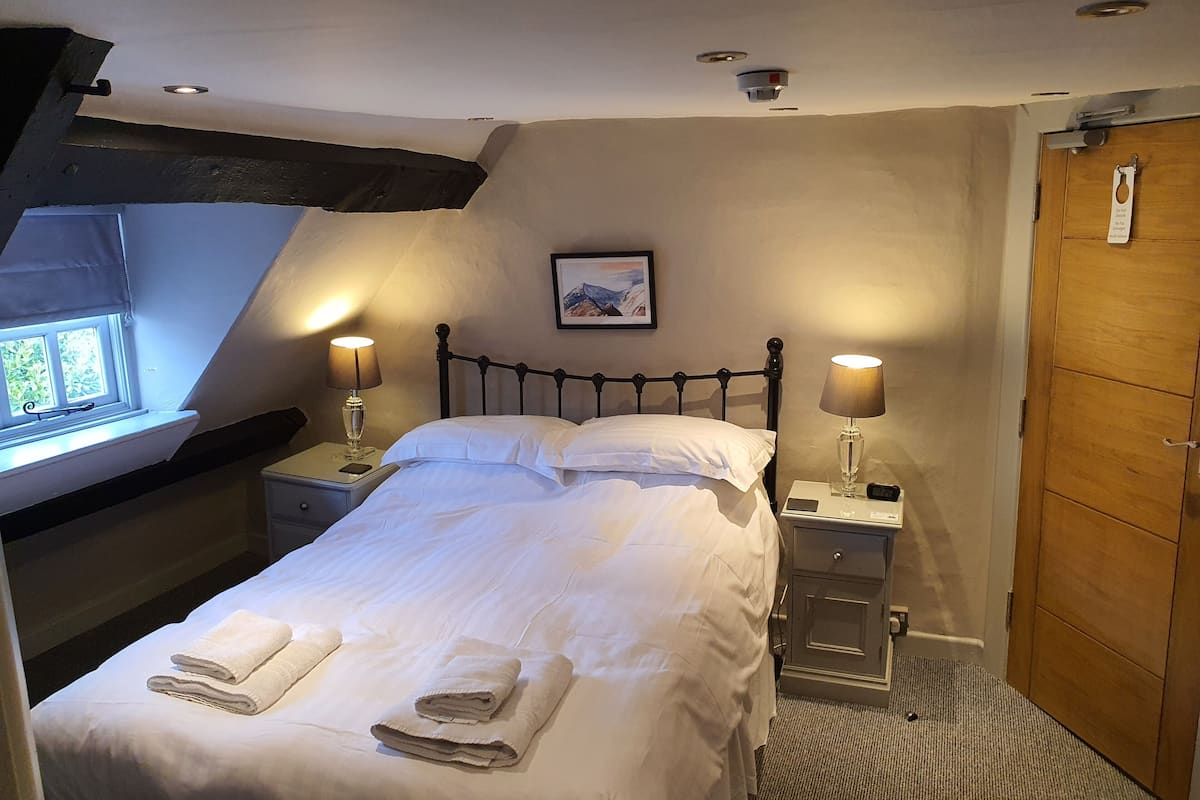 A neatly made bed with white linens and two folded towels, a bedside table with a lamp, a wooden wardrobe, and a framed picture on the wall.