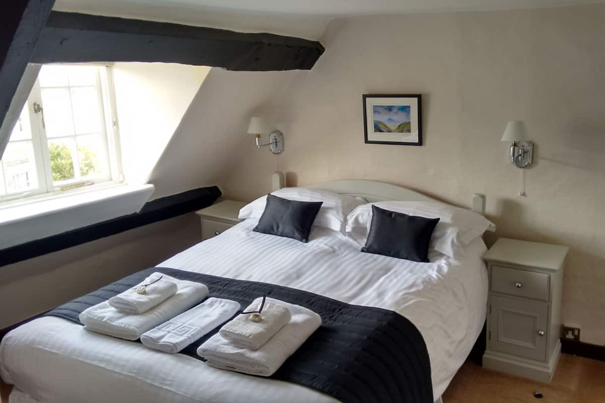 A neatly made bed with white and dark bedding, two bedside tables with lamps, a framed picture on the wall, and a window with a view of greenery.