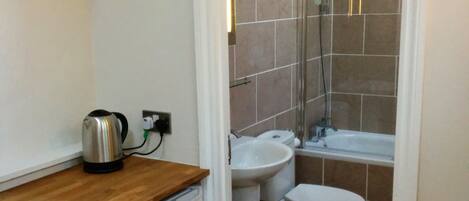 Un baño pequeño con ducha, inodoro, lavamanos y una kitchenette con hervidor.