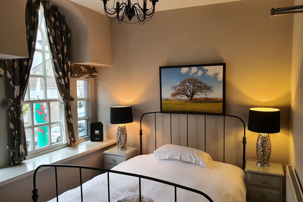 A bedroom with a bed, two bedside tables with lamps, a window with curtains, a chandelier, and a painting on the wall.