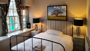 A bedroom with a bed, two bedside tables with lamps, a window with curtains, a chandelier, and a painting on the wall.