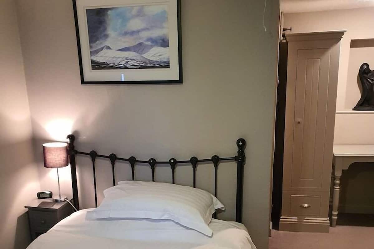 A neatly made bed with white linens and pillows, a bedside lamp, a framed picture on the wall, and a wooden wardrobe in the background.