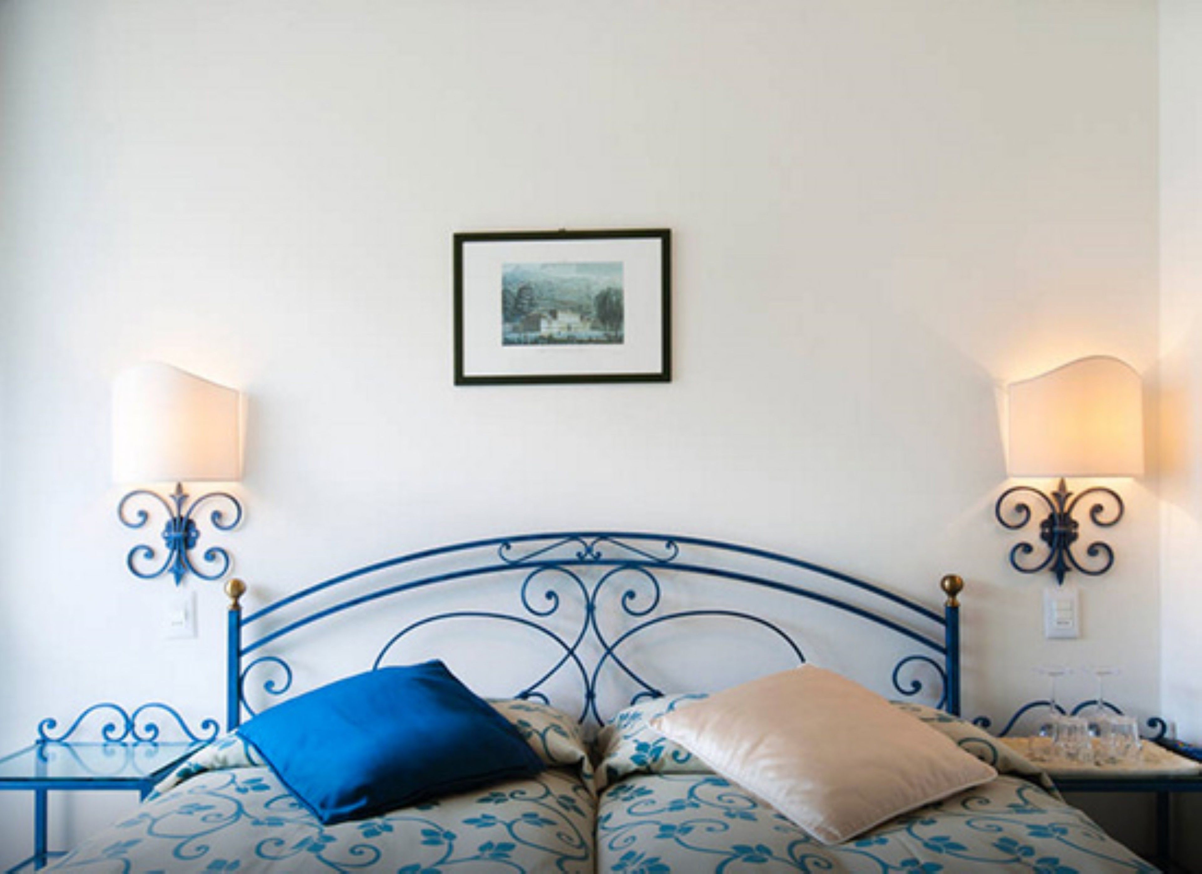 Una cama con estructura metálica, dos lámparas fijadas a la pared y un cuadro enmarcado en la pared.