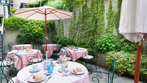 Een terras met ronde tafels, bedekt met roze tafelkleden, groene metalen stoelen en een grote parasol.