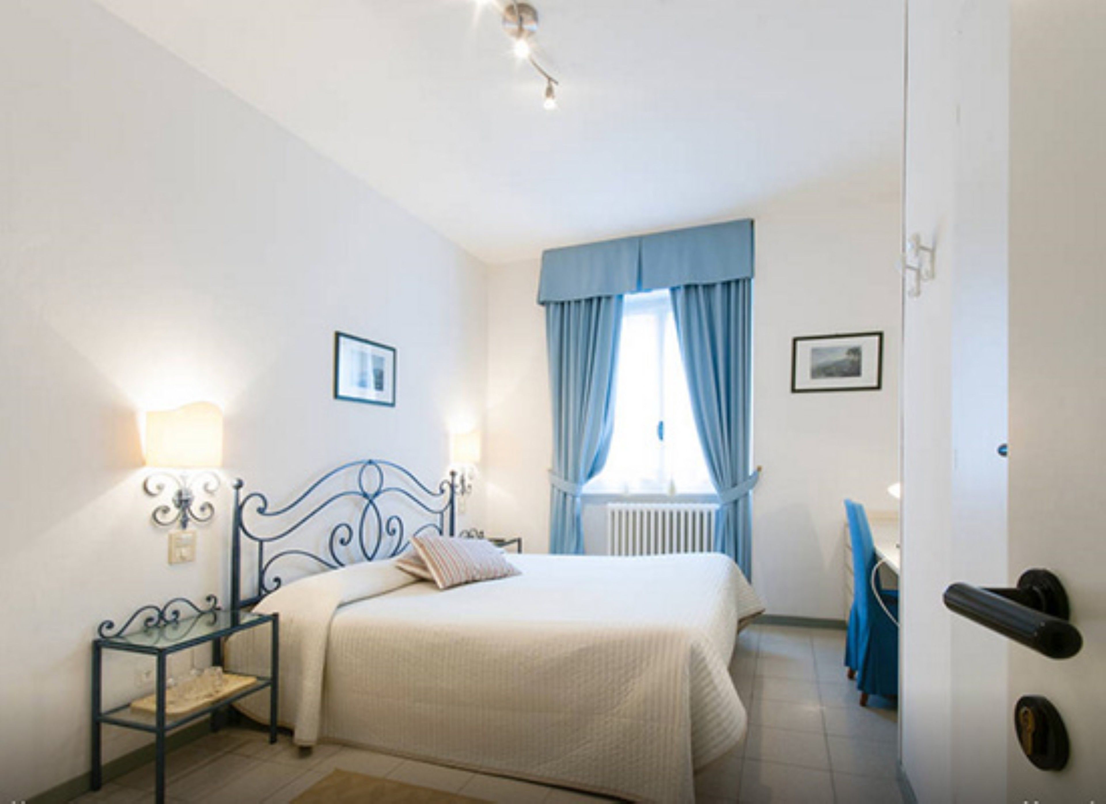 Een slaapkamer met een bed, nachtlampjes, een raam met gordijnen, een radiator en een deur met een handvat.