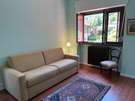 Ett vardagsrum med en beige soffa, en mönstrad matta, en trästol, ett element och ett fönster med utsikt över grönska och byggnader.