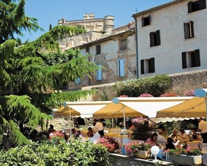 An outdoor café with tables and umbrellas, surrounded by greenery and historic buildings. - Les Géraniums (Le Barroux)