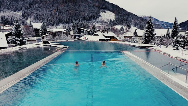 A swimming pool with two people in the water, surrounded by snow-covered trees and buildings. - Thermenwelt Hotel Pulverer (Bad Kleinkirchheim)