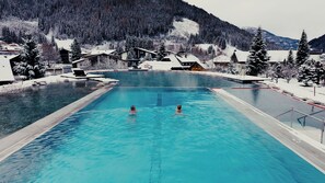 Una piscina con dos personas nadando, rodeada de árboles y edificios cubiertos de nieve.