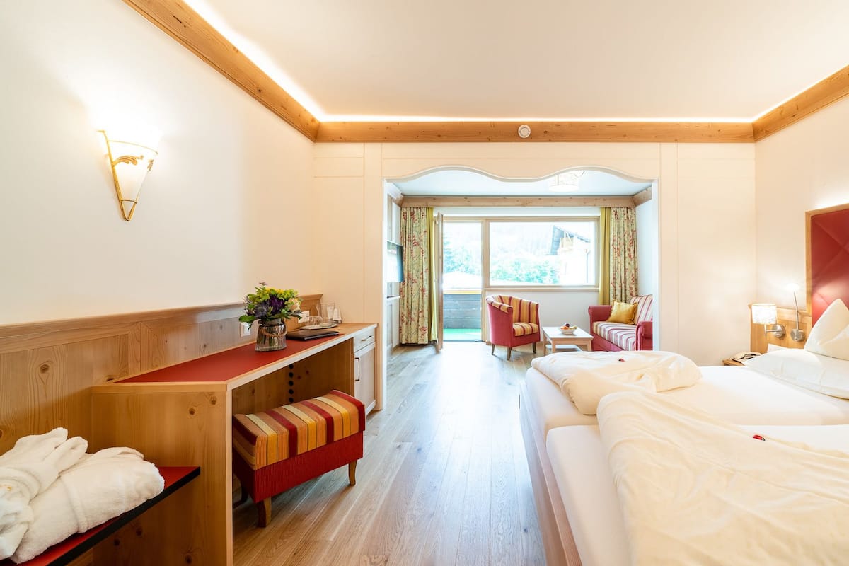 A hotel room with a wooden bench, a desk, a bed with red headboard, and a view of the garden through the window.