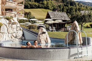A hot tub with two people, surrounded by rocks and umbrellas, with a mountain and wooden buildings in the background.