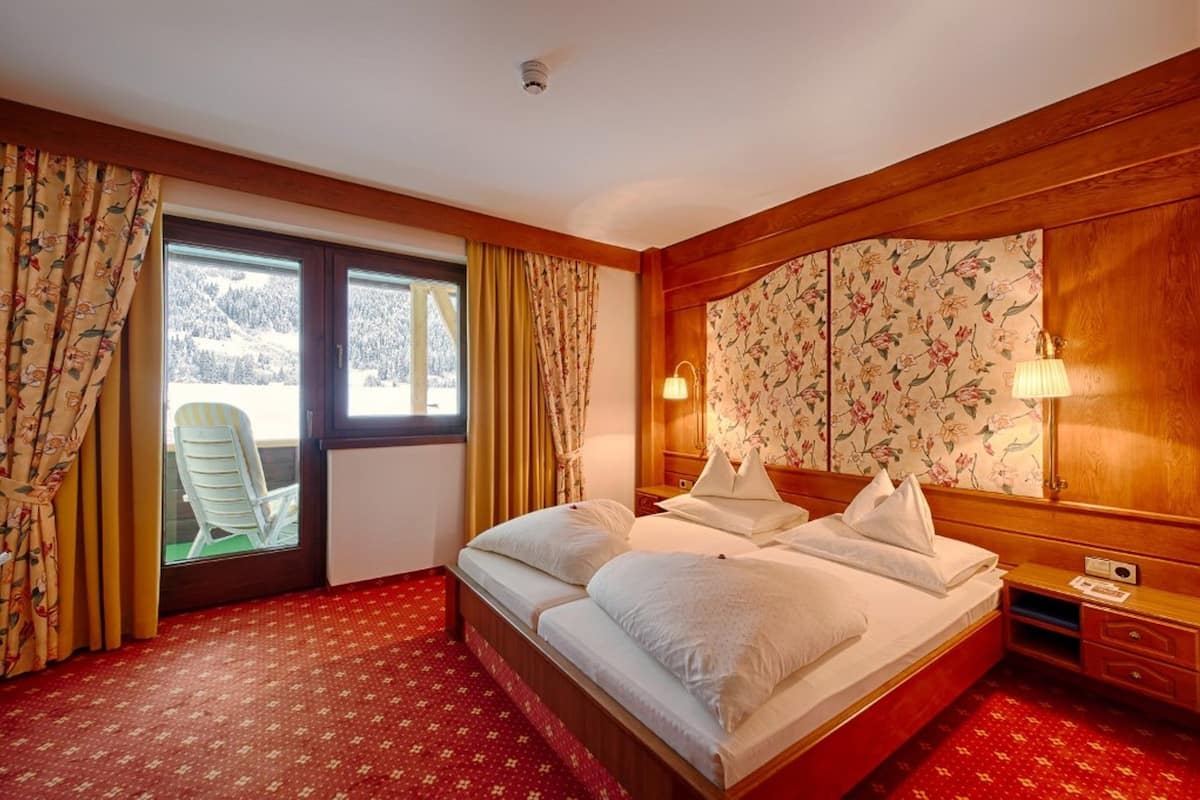 A hotel room with a bed, a nightstand, a lamp, a window with curtains, and a view of a snowy landscape.