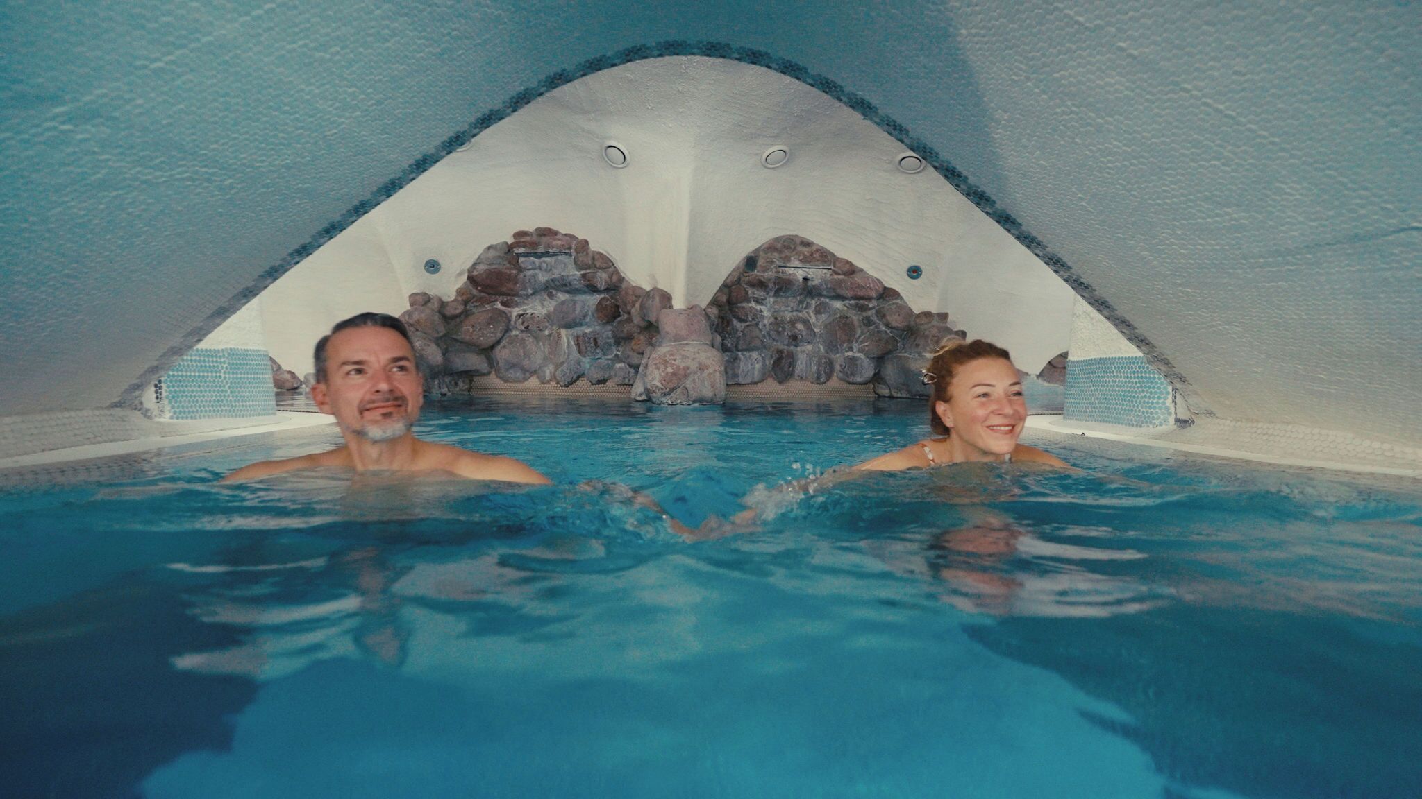 Un hombre y una mujer en un jacuzzi con un paisaje rocoso de fondo.