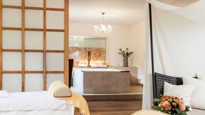 A modern hotel room with a wooden floor, a bed with white linens, a bedside table with a vase of flowers, and a large window with a view of the city. - Thermenwelt Hotel Pulverer (Bad Kleinkirchheim)