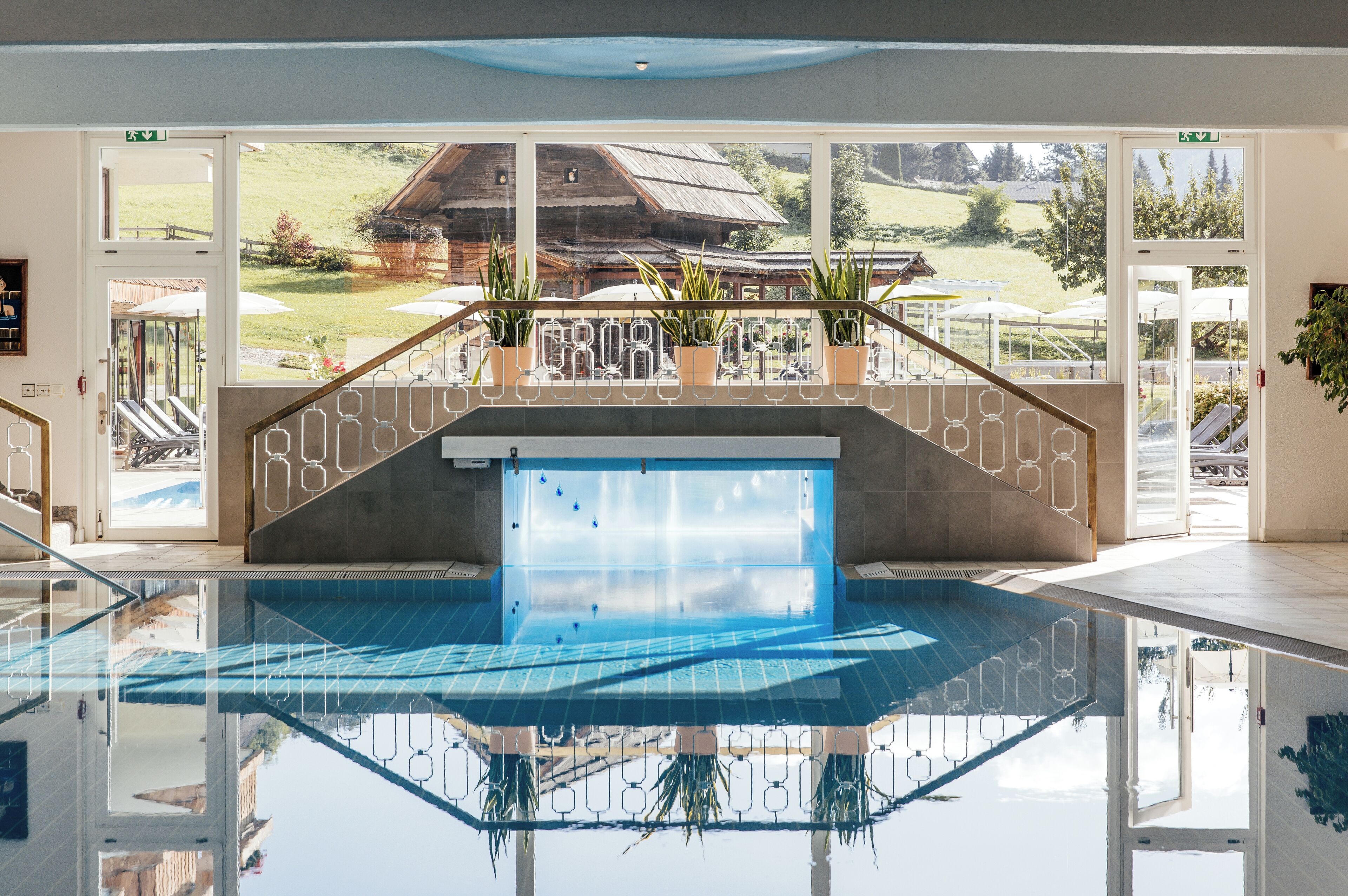 A modern indoor pool with clear glass walls, a wooden deck, and potted plants.