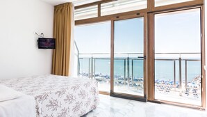 Habitación de hotel con una cama grande, un televisor montado en la pared y un balcón con vista a la playa y al océano.