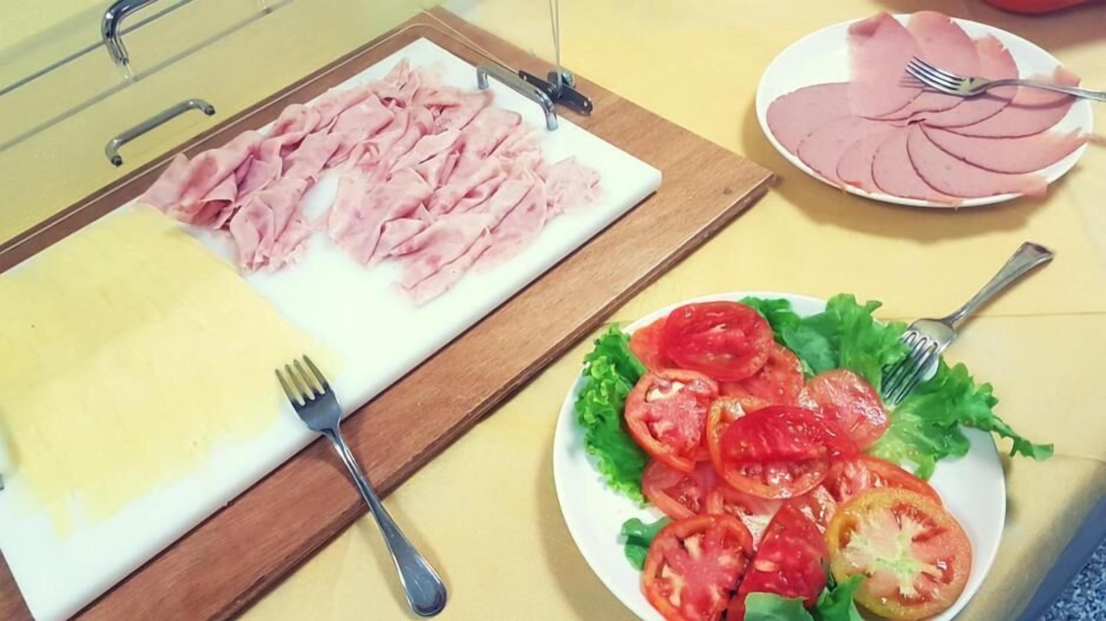 A cutting board with sliced ham and a plate of sliced tomatoes on a yellow table.