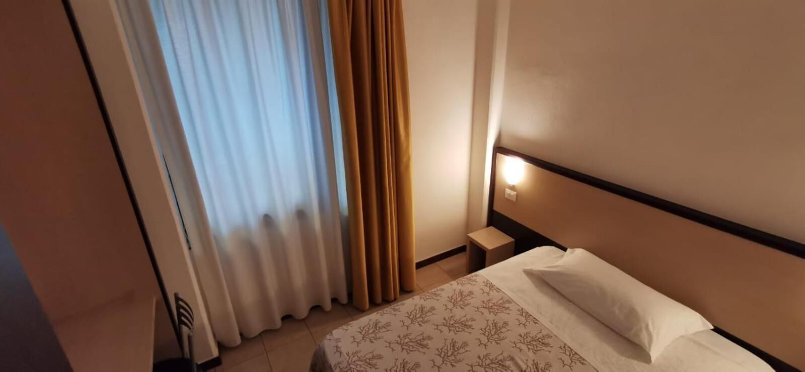 Una camera d'albergo con un letto, un comodino, una lampada a parete e una finestra con tende.