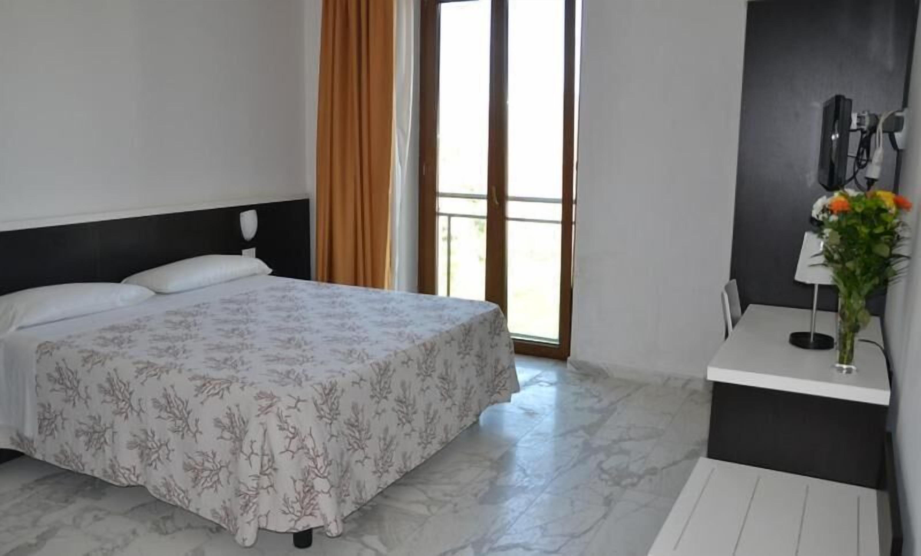 A hotel room with a bed, a window with curtains, a small table with a vase of flowers, and a wall-mounted lamp.