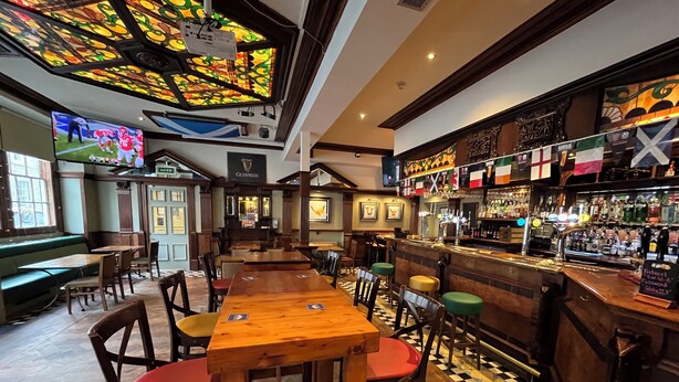 Una zona de bar con mesas y sillas de madera, una barra con diferentes botellas y un techo de vitrales.