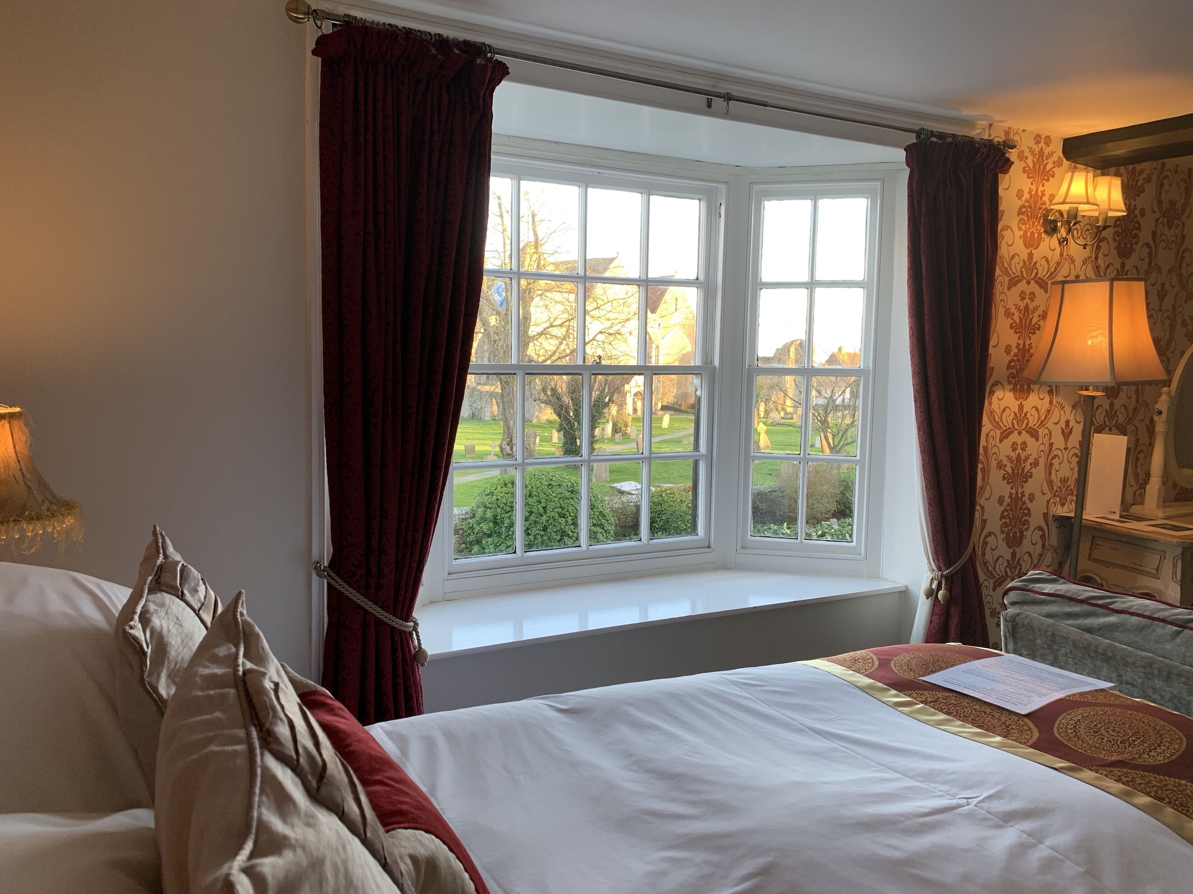 Een slaapkamer met een groot raam, een bed met witte beddengoed, een nachtkastje met een lamp, en uitzicht op een tuin.