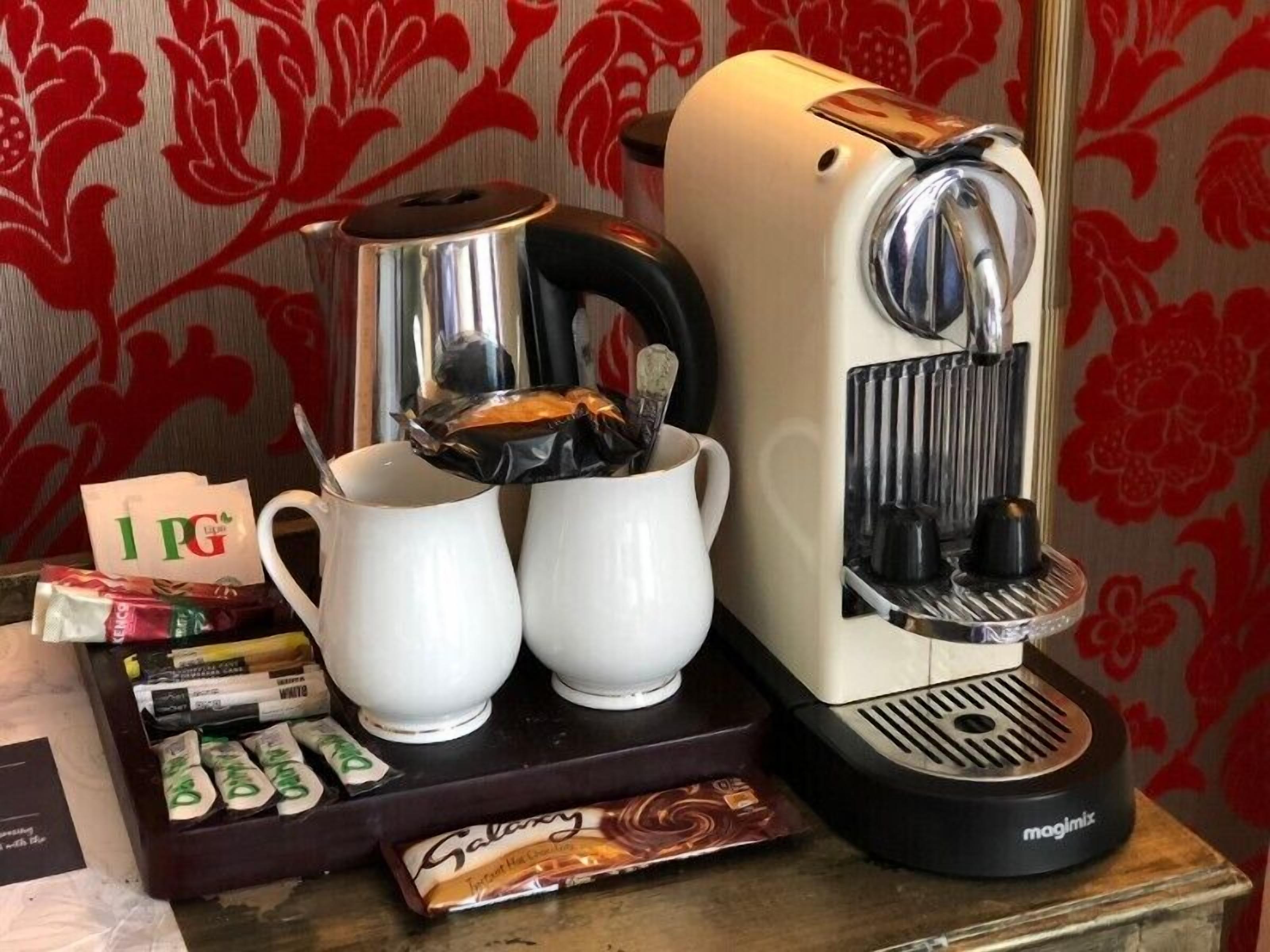 A coffee machine with two white cups, a tray with coffee pods, and a packet of Galantine chocolate.