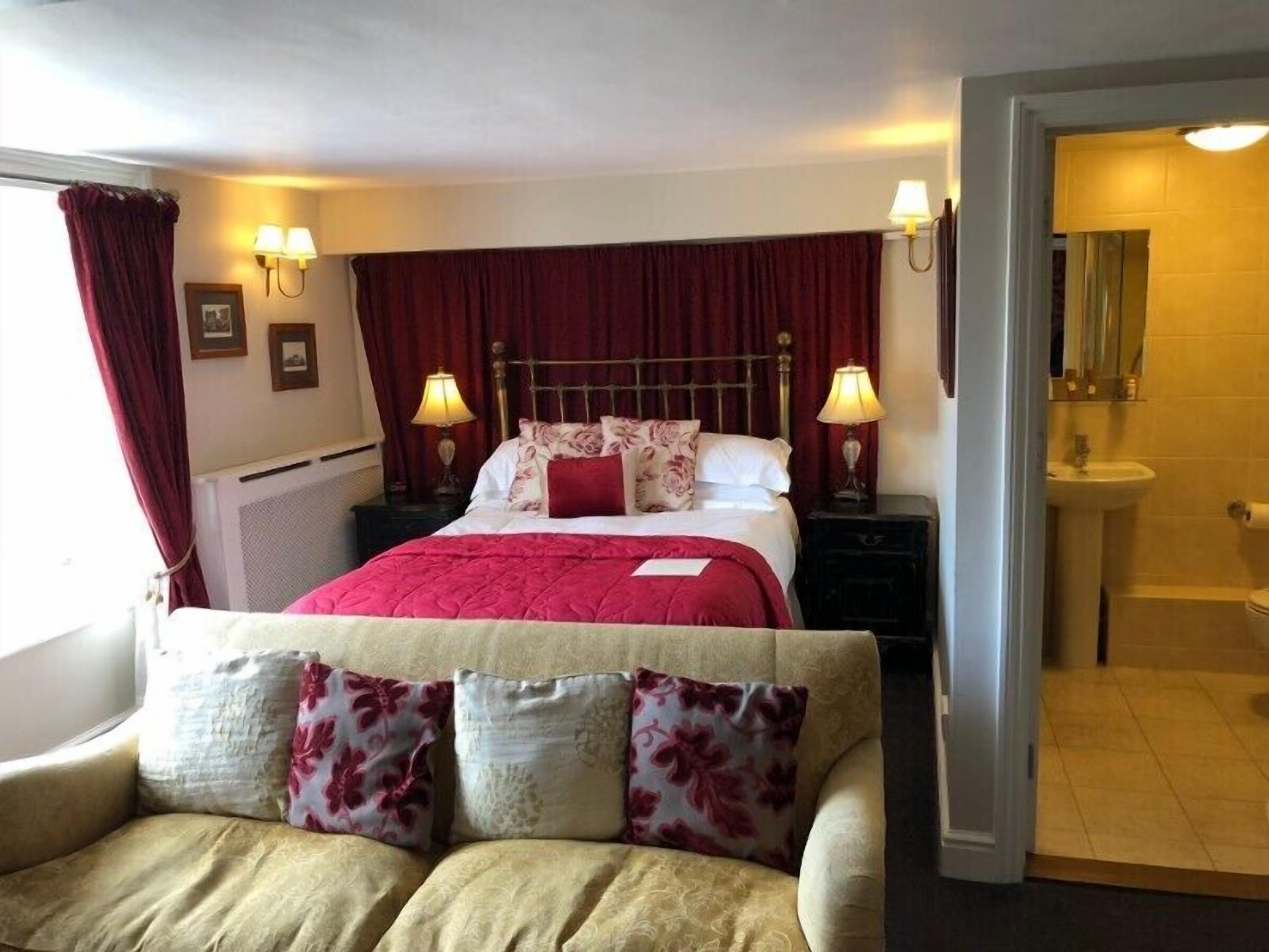 Ein Hotelzimmer mit einem Bett, zwei Nachttischlampen, einem Nachttisch, einer Couch mit floralen Kissen und einem durch eine offene Tür einsehbaren Badezimmer.
