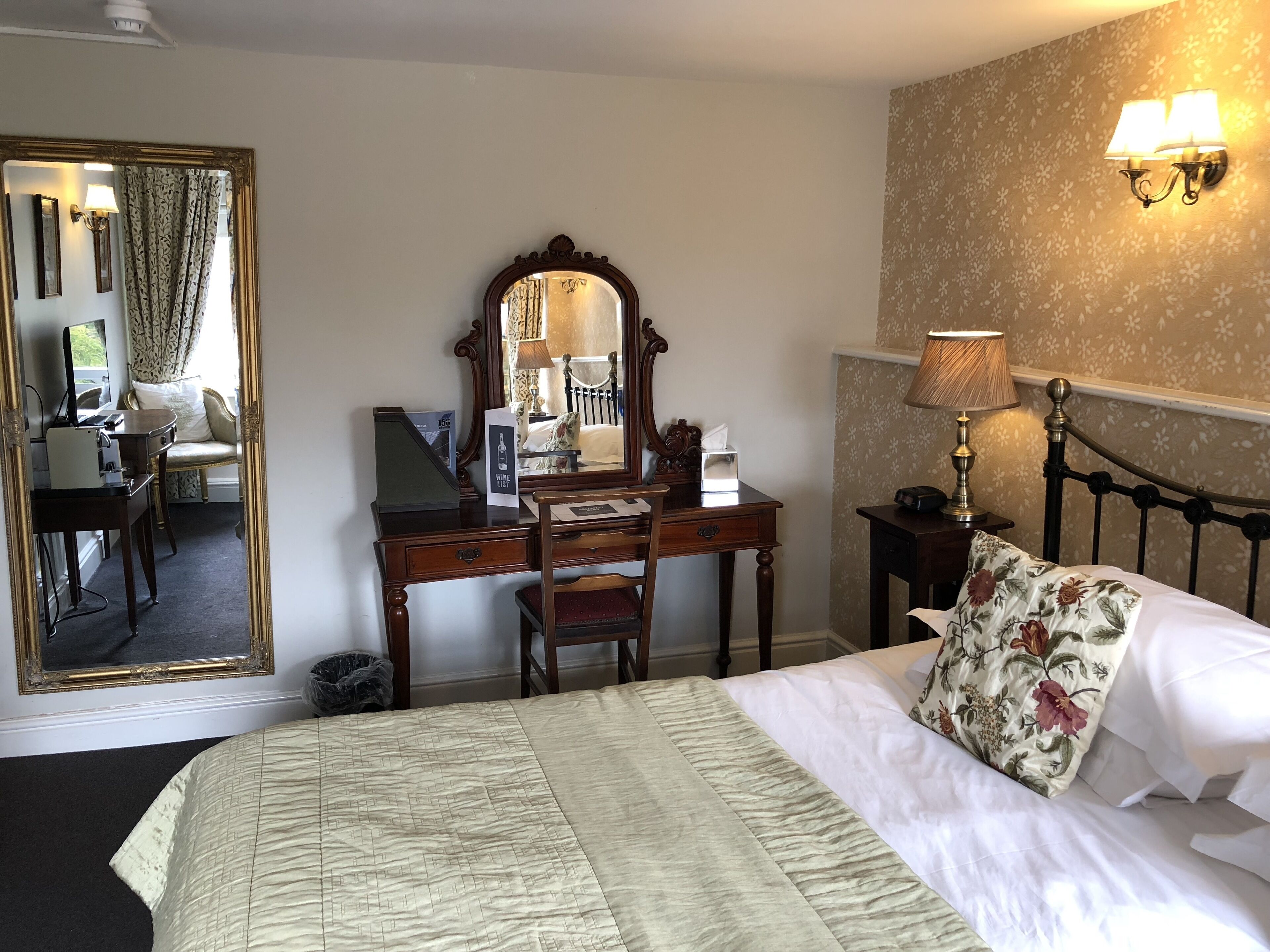 A hotel room with a large mirror, a desk with a computer, a bed with floral bedding, and a wall-mounted lamp.