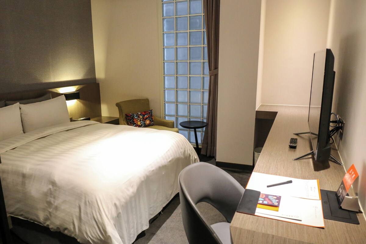 Une chambre d’hôtel comprenant un lit, un bureau avec un ordinateur, une chaise, une petite table et une fenêtre avec des rideaux.