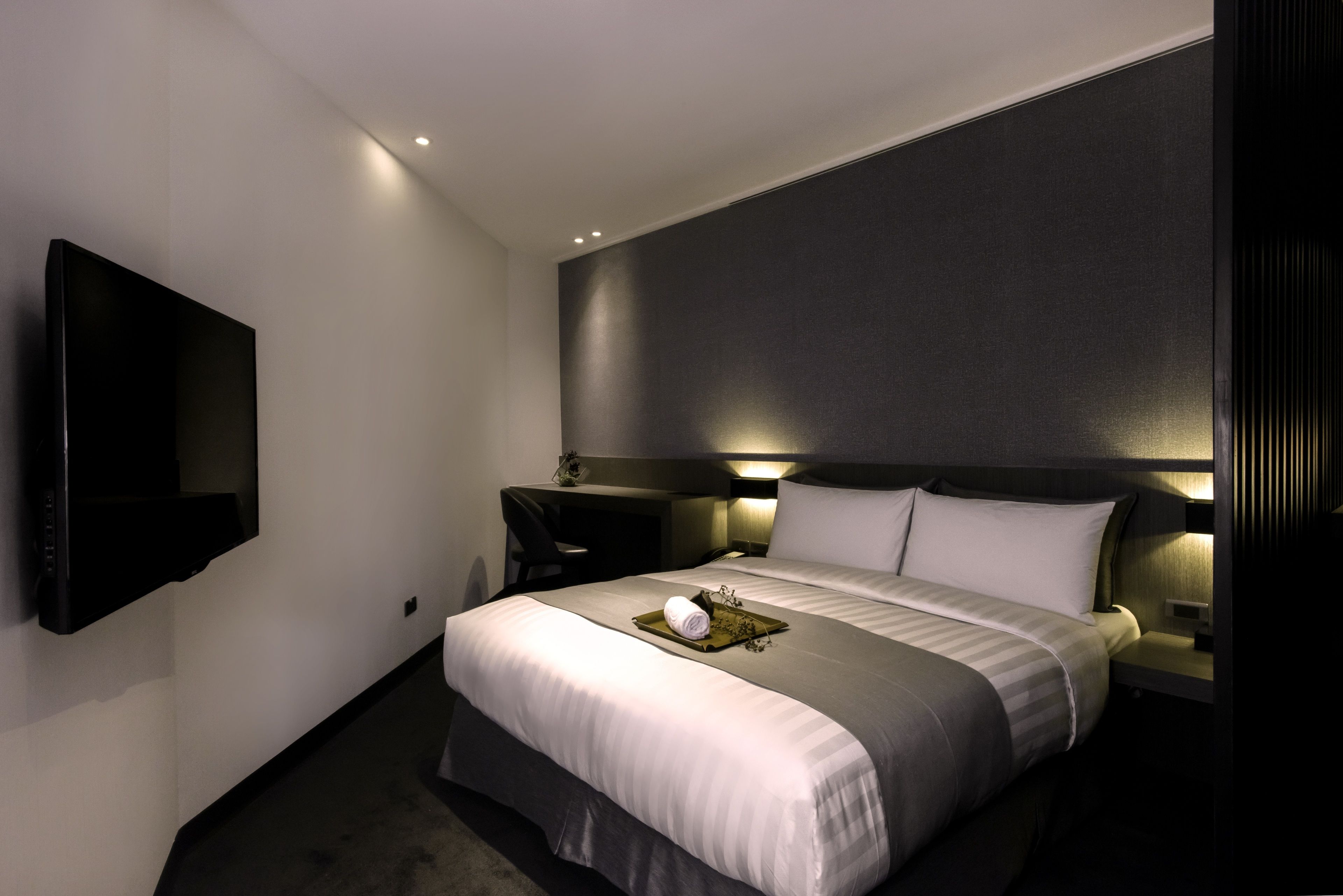 Une chambre d’hôtel moderne dotée d’un grand lit, d’un téléviseur à écran plat fixé au mur, d’un bureau et d’une table de chevet avec une lampe.