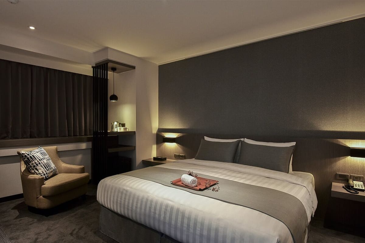 Une chambre d’hôtel moderne avec un grand lit, une chaise, des tables de chevet avec des lampes et une fenêtre avec des rideaux.