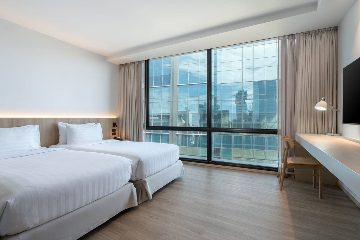 A hotel room with two beds, a desk, a chair, a large window with curtains, and a flat-screen TV.