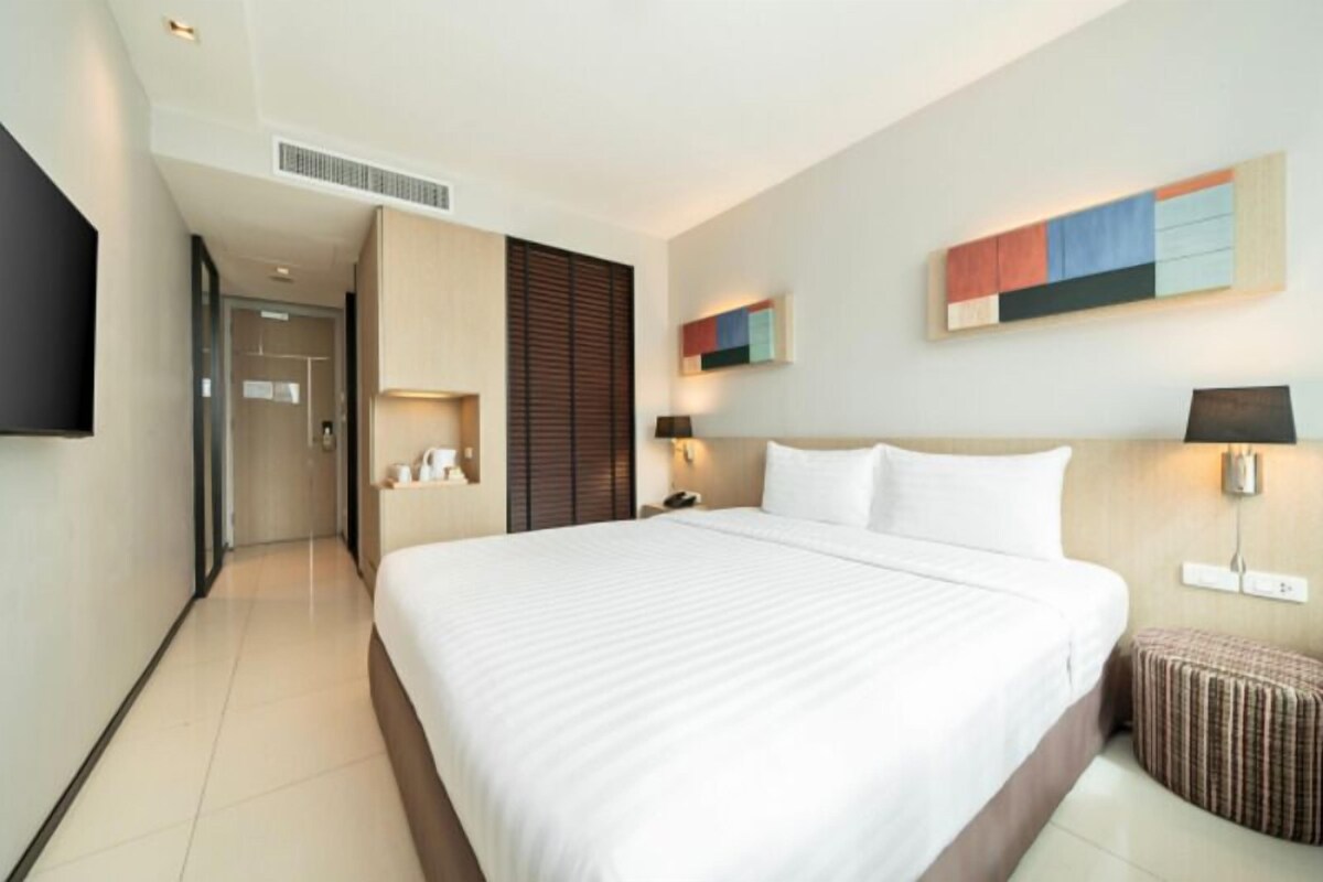 A hotel room with a large bed, a bedside table, a lamp, a wall-mounted TV, and a small ottoman.