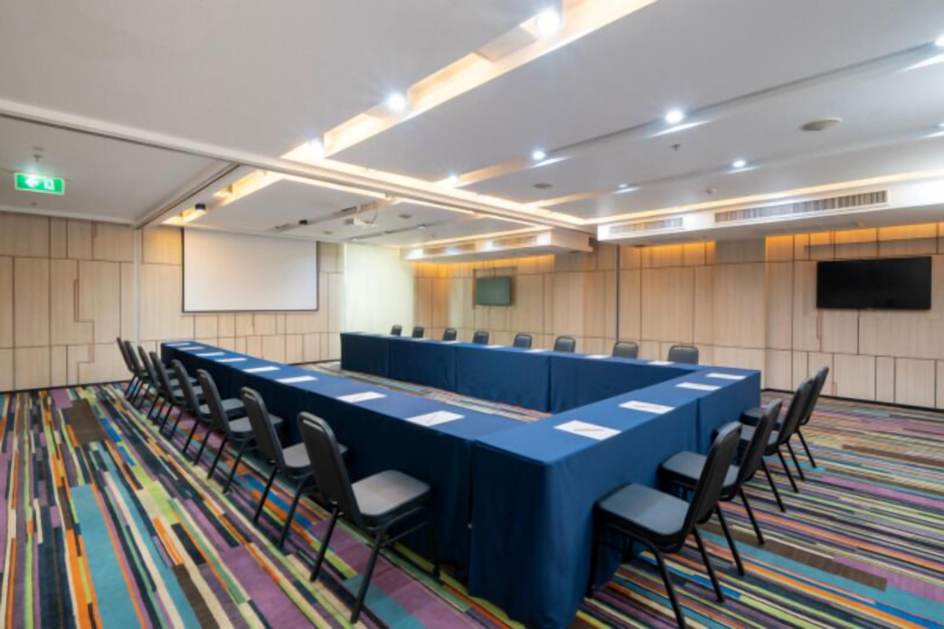 Une salle de conférence avec une longue table, des chaises, un écran de projection et un téléviseur fixé au mur.