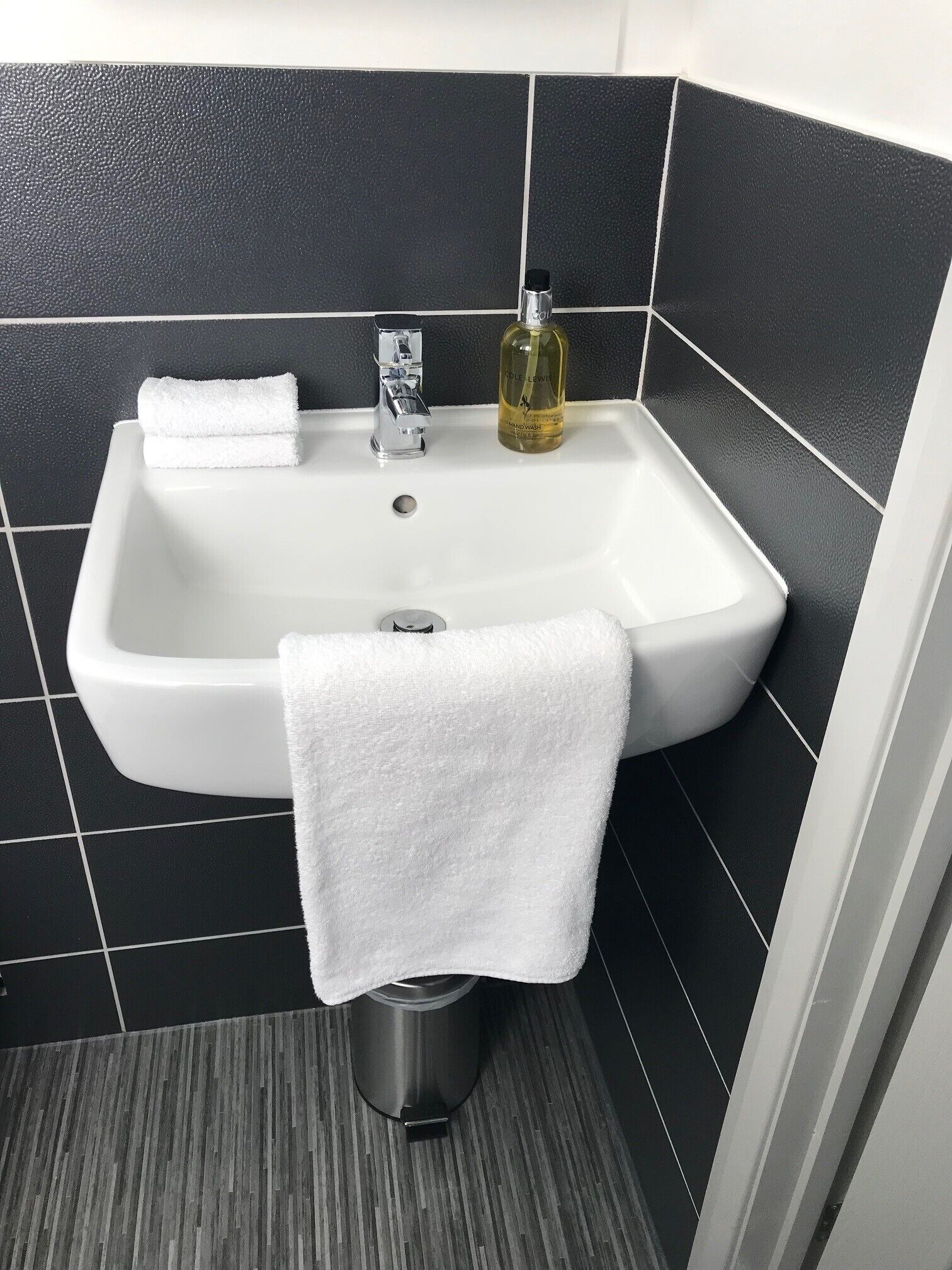 Une salle de bain avec un lavabo blanc, des carreaux noirs, un flacon de savon liquide et une serviette blanche posée sur un porte-serviettes en métal.